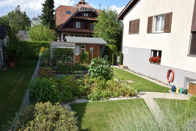 Well-maintained garden with lawn, flower beds, and a small wooden shed. The garden is bordered by a white fence and features a variety of plants and shrubs. A house with a red-tiled roof and white walls is visible in the background.