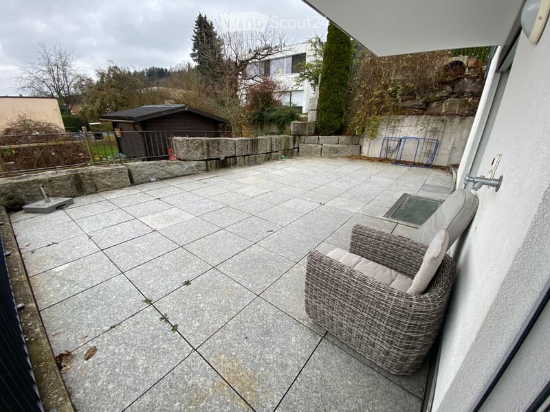 Balcony with stone tiles, a wicker chair, and a small table. The area is enclosed by a stone wall and has a view of the surrounding greenery.