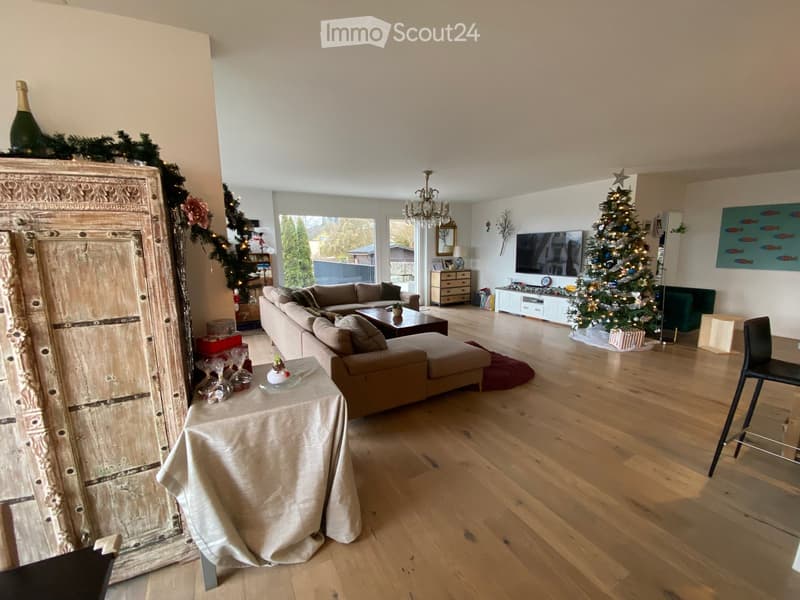 Spacious living room with wooden flooring, a large sectional sofa, coffee table, ottoman, TV, and two Christmas trees. A large cabinet and a small table with a decorative centerpiece are also present.