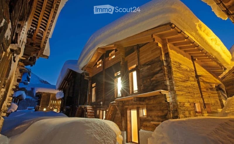 A large wooden building with snow-covered roof and walls, illuminated by warm lights, typical of a mountain lodge or chalet.