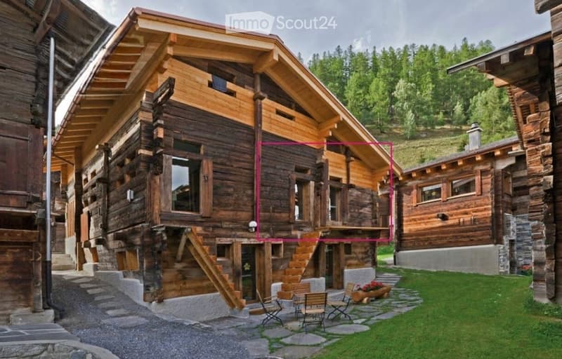 A large wooden chalet with multiple stories, large windows, and a stone base, surrounded by greenery and a small patio area with outdoor furniture.