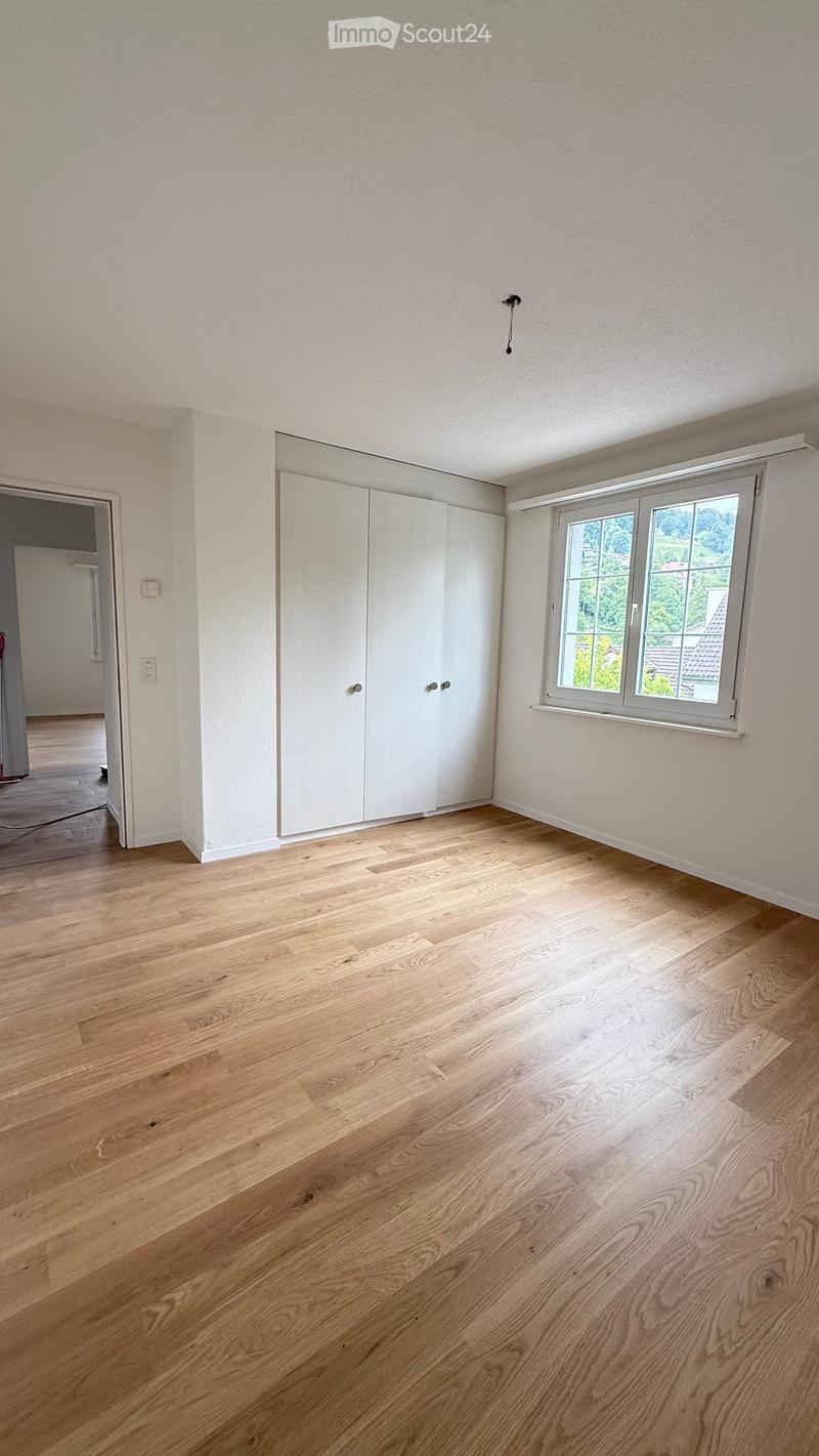 Empty room with light wooden flooring, white walls, a large window with a view, and a white closet with double doors.