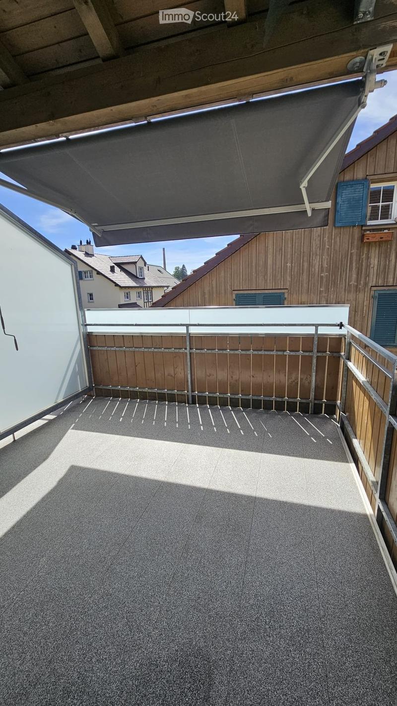 Roof terrace with a modern metal railing, wooden privacy screen, and a retractable awning. The floor is covered with a textured gray material, and the surrounding area includes wooden walls and neighboring buildings.