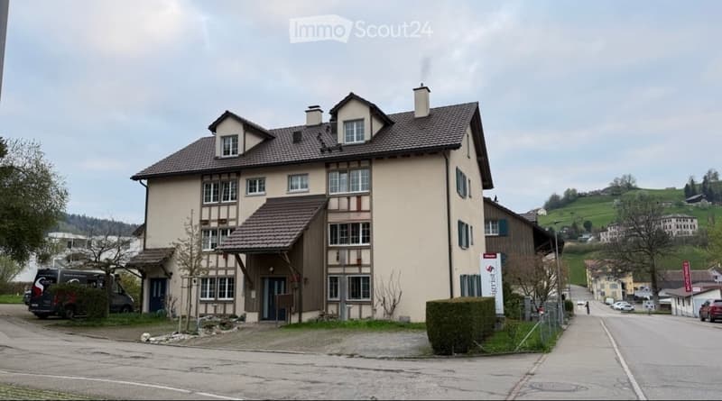A multi-story residential building with a beige facade, brown roof, and multiple windows. The building has a covered entrance and is situated in a suburban area with trees and other houses visible in the background.