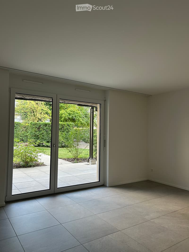 Empty room with large sliding glass doors leading to a garden, tiled floor, and neutral-colored walls.