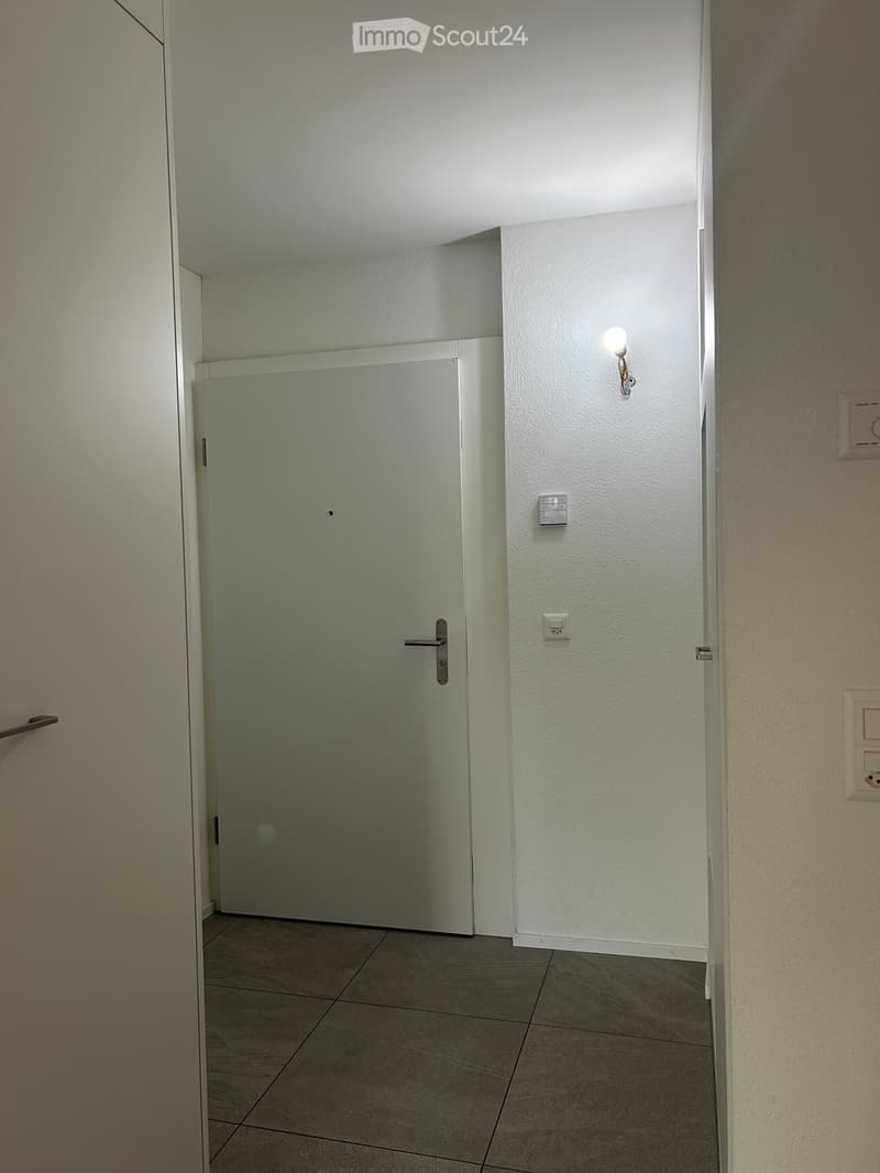 Empty hallway with tiled floor, white walls, and a closed door. Light fixture mounted on the wall and electrical outlets visible.