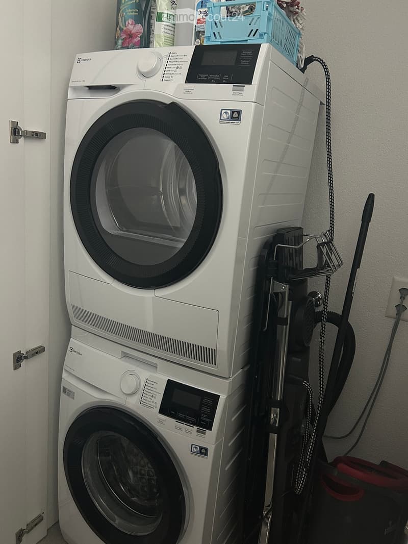 A compact bathroom with a stacked washing machine and dishwasher unit, a vacuum cleaner on a stand, and cleaning supplies on top.