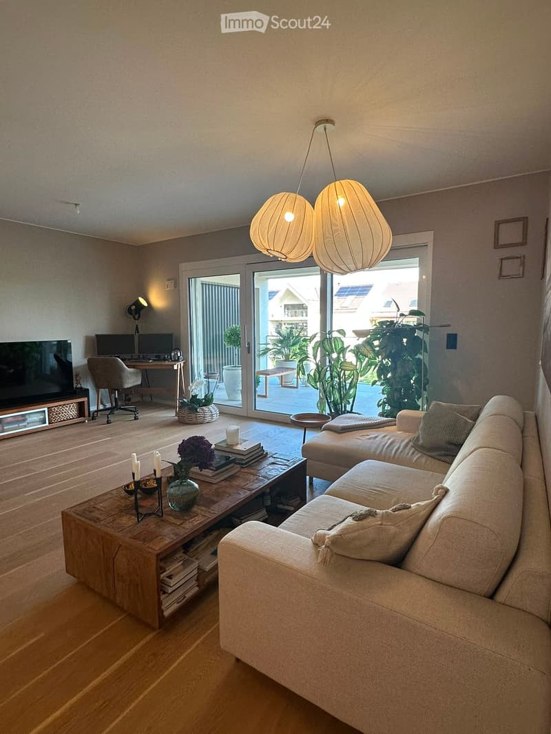 Modern living room with light wooden flooring, a large sofa, coffee table, desk with computer, and a view through large glass doors.