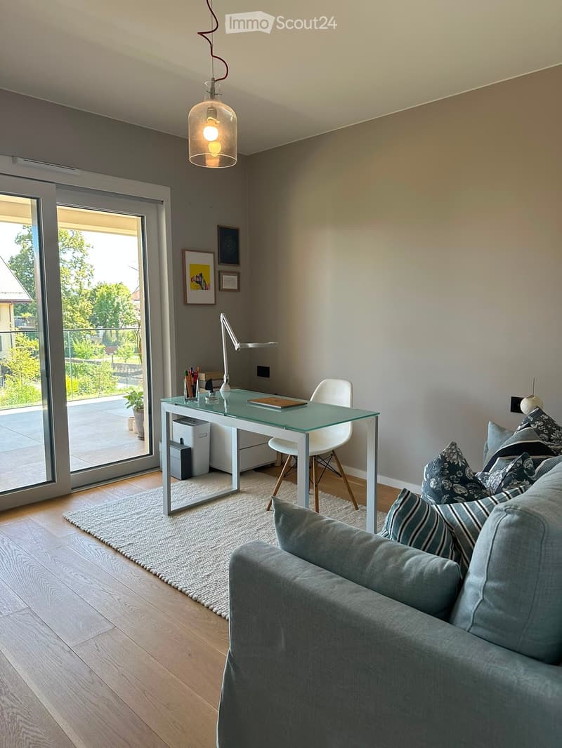 Modern living room with a grey sofa, coffee table, desk, and large windows leading to a balcony.