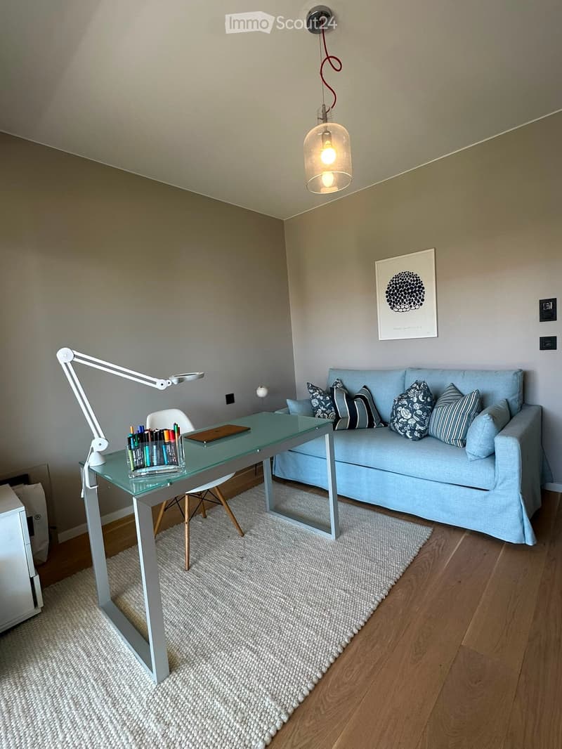 A modern, well-lit room with a light blue sofa, a glass desk, a white adjustable lamp, and a white chair. The room features a white shag rug and a framed picture on the wall.