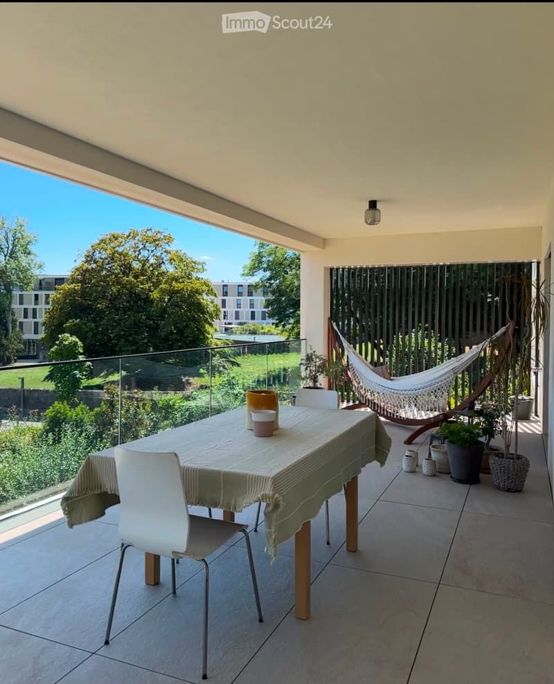 Balcony with a dining table, chairs, a hammock, and potted plants, overlooking a green area with trees and a building in the background.
