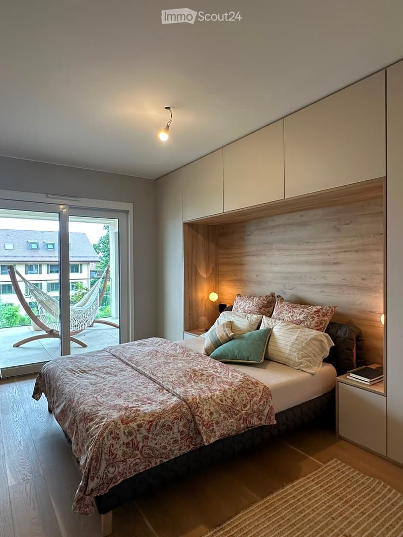 Modern bedroom with a large bed, patterned duvet, multiple pillows, nightstand with a book, and a built-in wooden headboard. The room features wooden flooring and a large glass door leading to a balcony with outdoor furniture visible.