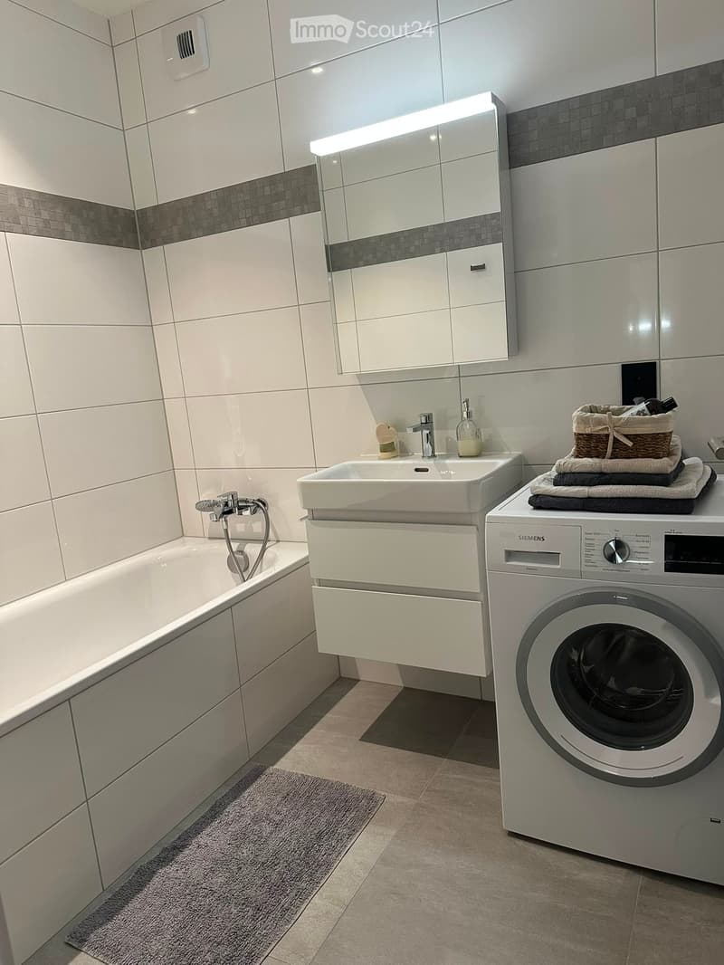Modern bathroom with white tiles, bathtub, sink, mirror, and washing machine. Includes towels and a basket on top of the washing machine.