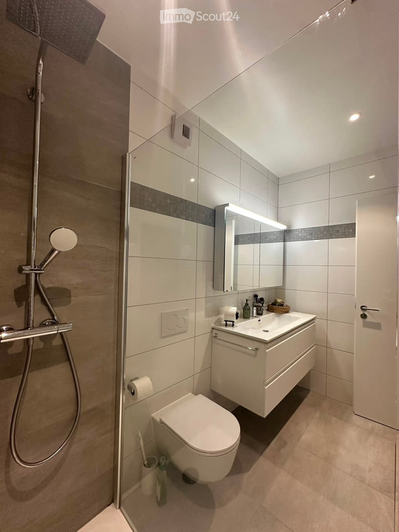 Modern bathroom with white tiles, a toilet, a sink with a mirror, a shower, and a small basket on the sink counter.
