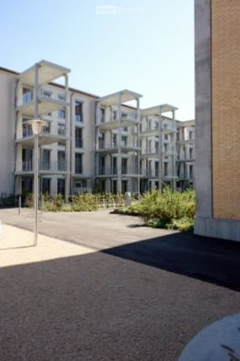 Modern multi-story residential building with balconies and contemporary design, surrounded by greenery and paved area.