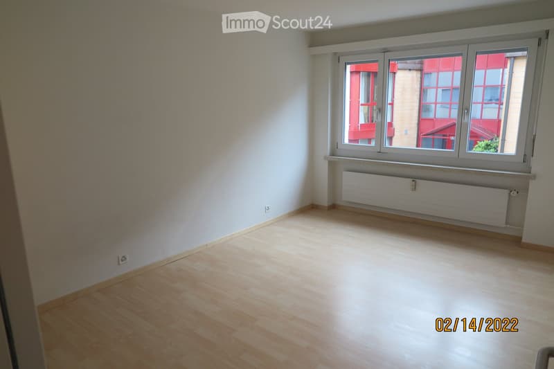 Empty room with light wooden flooring, white walls, and large windows with white frames. A radiator is visible beneath the windows.