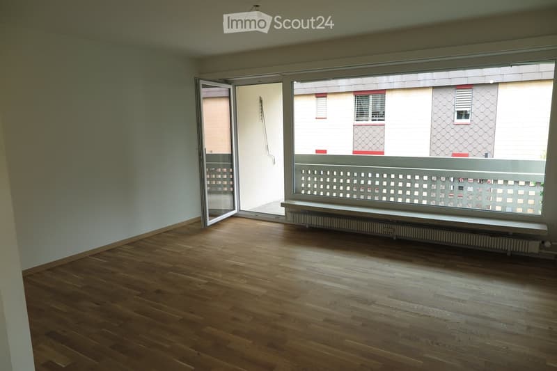 Empty room with wooden flooring, large window with a decorative white lattice, and a radiator below the window. The room has white walls and a sliding glass door leading to a balcony.