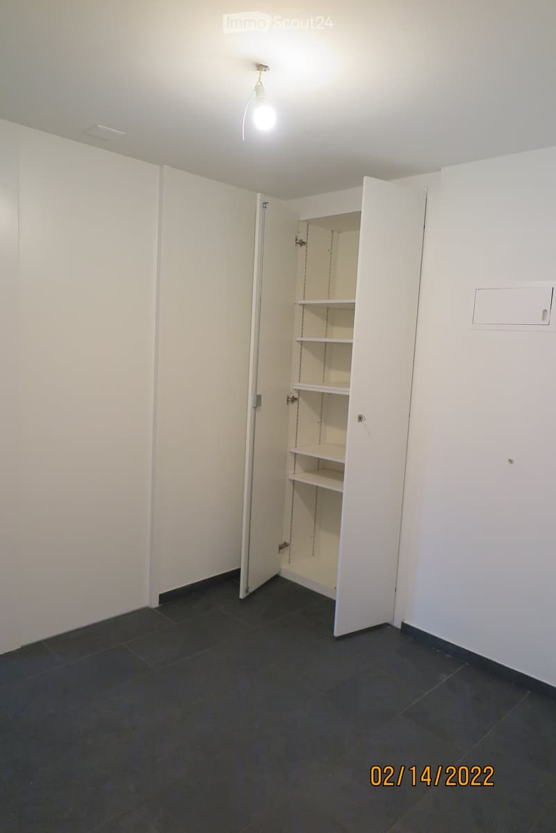 Empty room with white walls, dark tiled floor, and an open white cabinet with empty shelves.