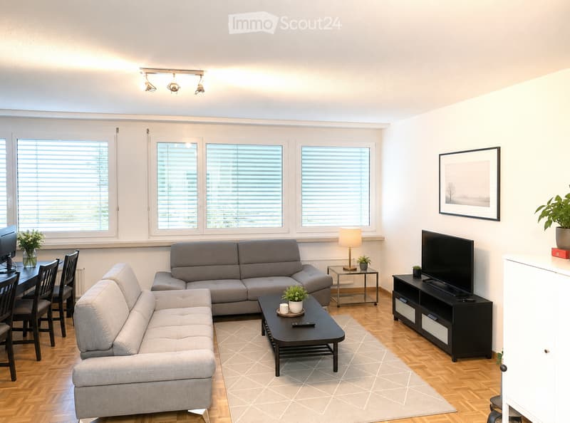 Modern living room with light-colored walls, wooden flooring, large windows with blinds, a sectional sofa, coffee table, dining table with chairs, TV stand, and decorative plants.