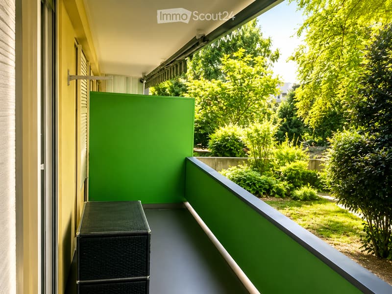 Balcony with green railings, black wicker storage box, and a view of a lush garden with trees and shrubs.