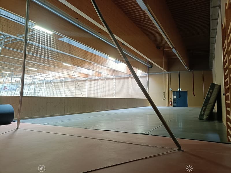 Large, open gymnasium with wooden beams, large windows, and a padded floor. No furniture or equipment is visible.