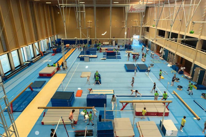 A spacious indoor gymnastics training facility with multiple padded exercise areas, balance beams, uneven bars, and a vaulting table. Young gymnasts and coaches are practicing various routines.