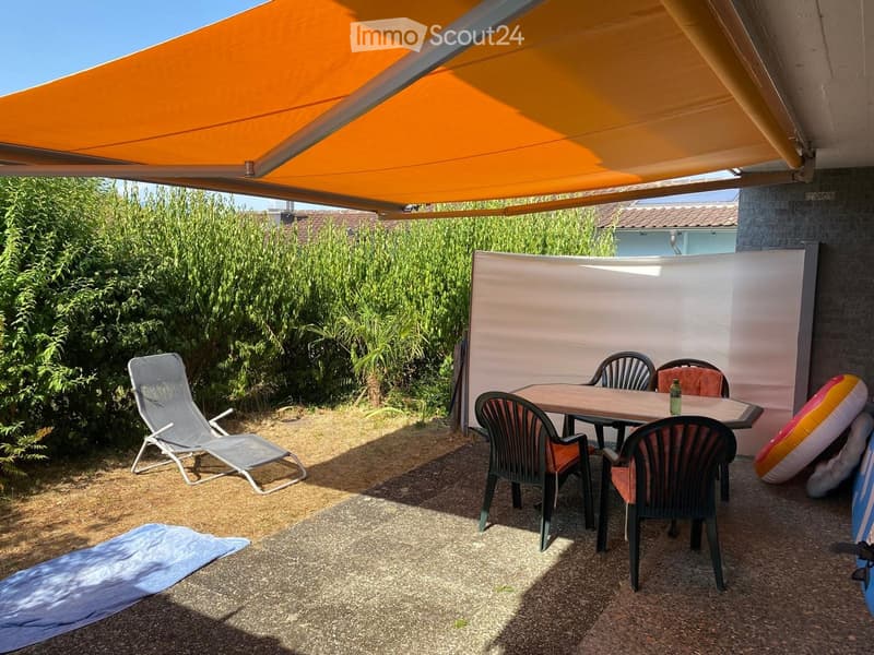 Outdoor patio with a table, chairs, a lounge chair, and a blue towel on the ground, surrounded by greenery and an orange awning.