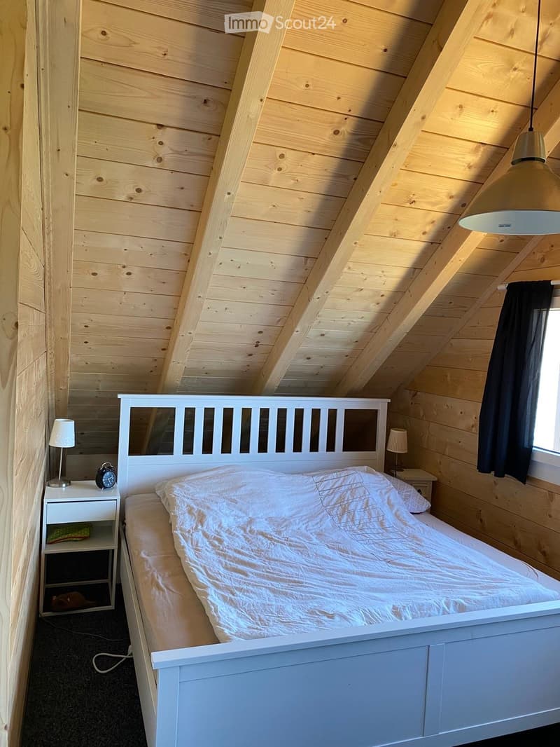 Cozy bedroom with wooden walls and slanted ceiling, featuring a white bed with a headboard, nightstands with lamps, and a clock on one side.
