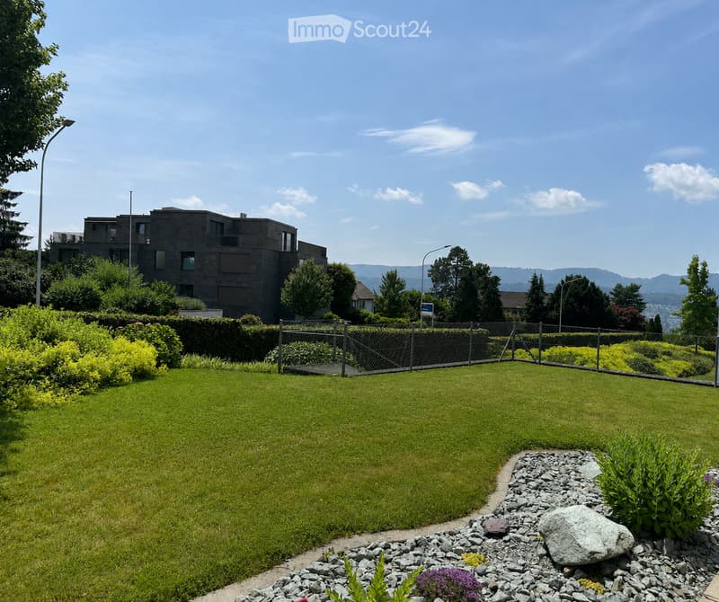 Well-maintained garden with a large lawn, rock border, and surrounding greenery. A modern building with balconies is visible in the background.