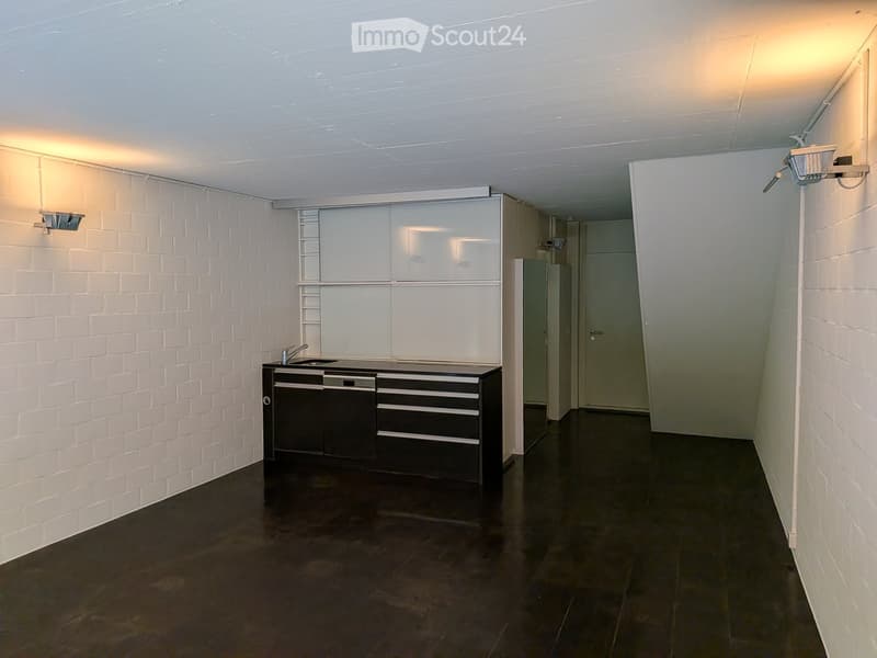 Empty room with white brick walls, dark floor, and a small kitchen area with cabinets and sink.