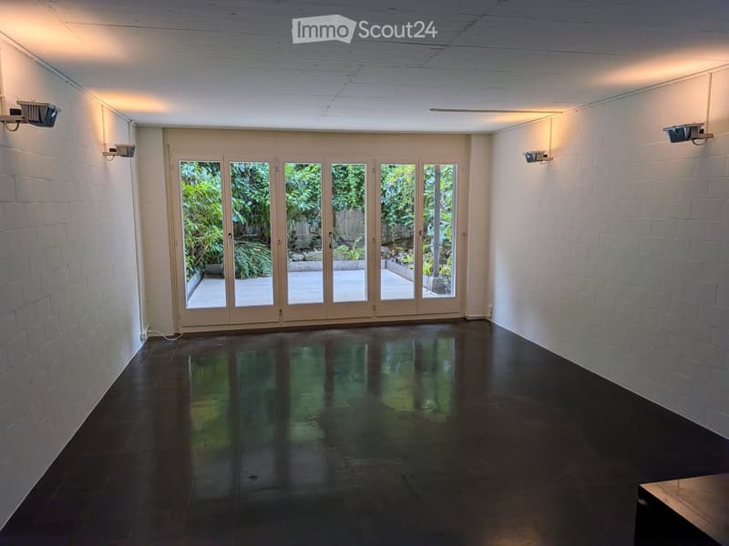 Empty room with white brick walls, dark polished floor, large glass doors leading to a garden, and recessed lighting.