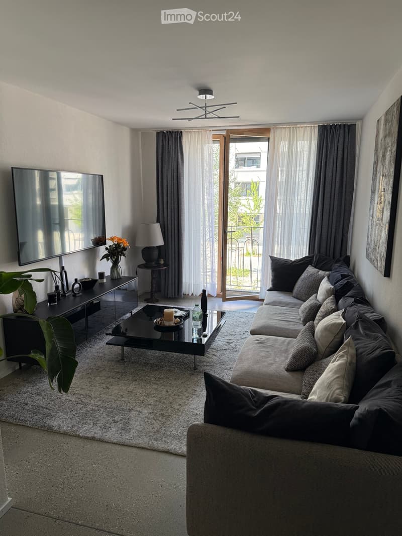 Modern living room with a large sectional sofa, coffee table, side table with lamp, television mounted on the wall, and a view to a balcony through large windows.
