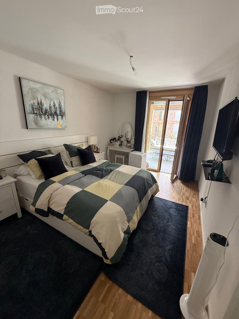 A modern bedroom with a large bed, nightstand, mirror, and a TV mounted on the wall. The room features wooden flooring, a dark rug, and a sliding glass door leading to a balcony.