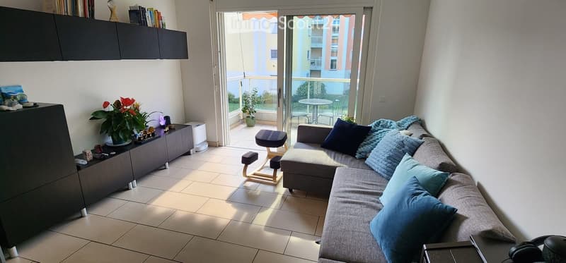 Modern living room with a grey sectional sofa, blue throw pillows, a small wooden stool, a black cabinet with books and decorative items, and a view of a balcony with outdoor furniture.