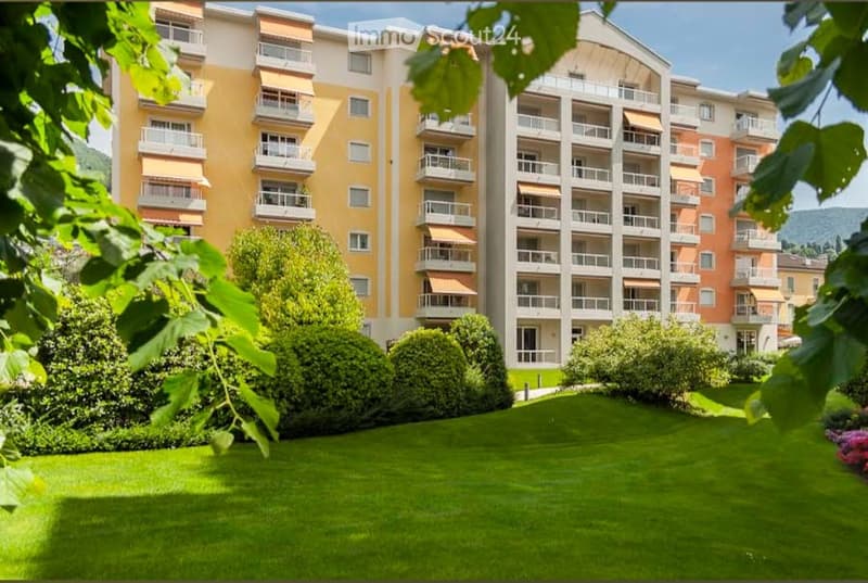 Multi-story residential building with balconies, surrounded by a well-maintained lawn and trees.