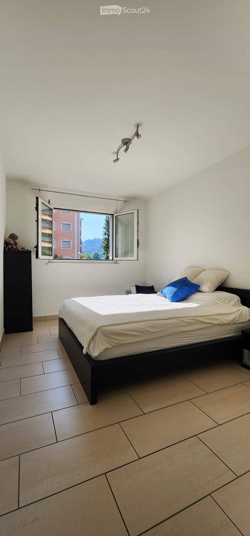 Modern bedroom with a large bed, white bedding, blue pillows, a nightstand, and a wardrobe with toys on top.