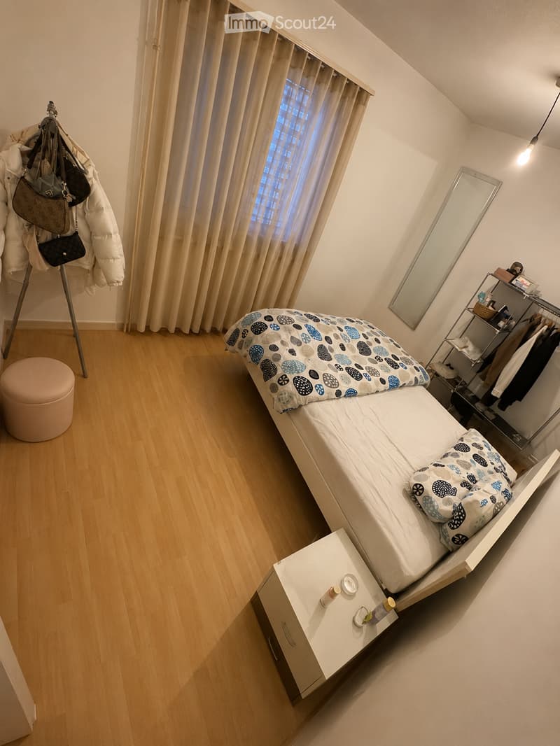 Cozy bedroom with a single bed, nightstand, stool, mirror, and clothing rack. The room features wooden flooring and large windows with curtains.