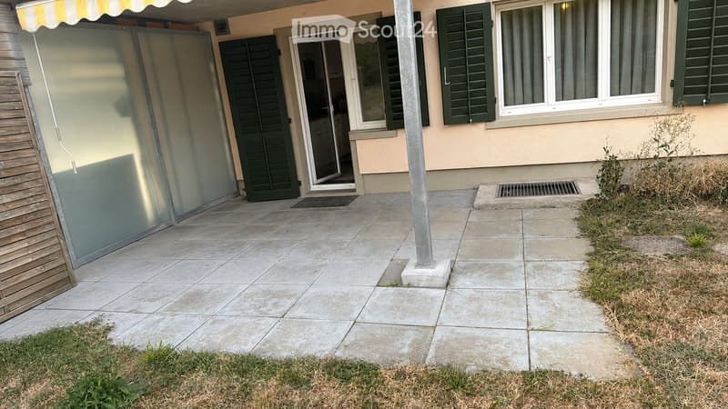 Patio area with tiled flooring, green shutters, and a small grassy area.
