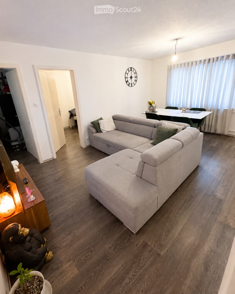Modern living room with a grey sectional sofa, wooden coffee table, dining area with chairs and table, wall-mounted clock, and decorative items including a plant and a lamp.