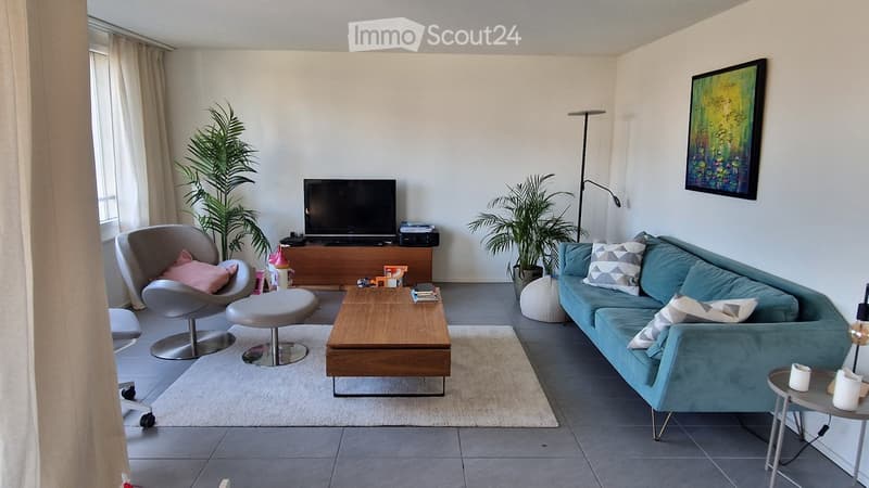 Modern living room with a grey sofa, wooden coffee table, grey chair, and television on a wooden stand. The room features large windows with curtains, a floor lamp, and decorative plants.