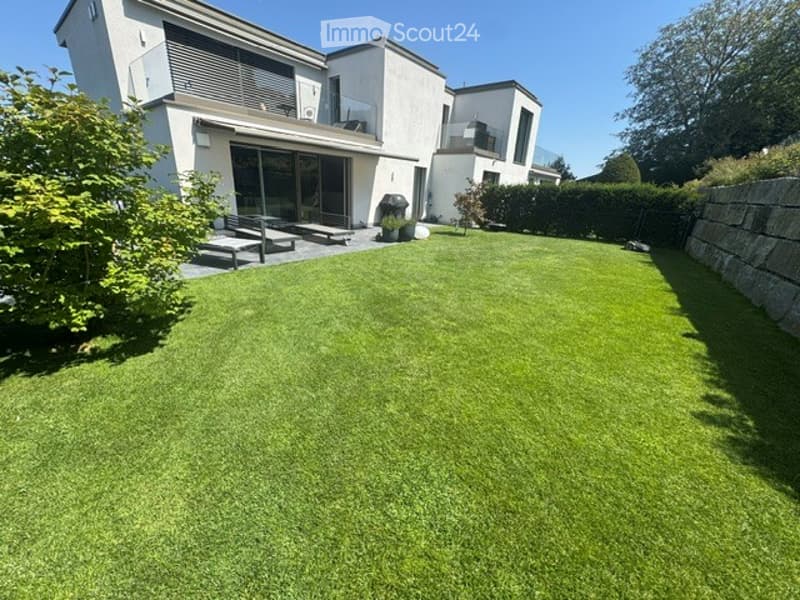 Well-maintained lawn in front of a modern house with large windows and a balcony, surrounded by greenery and a stone wall.