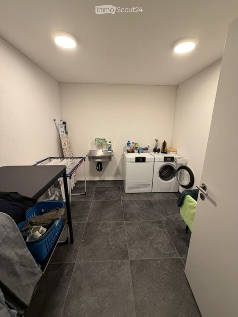 Laundry room with a washing machine, dryer, small table, chair, and a sink. The room has a tiled floor and white walls.