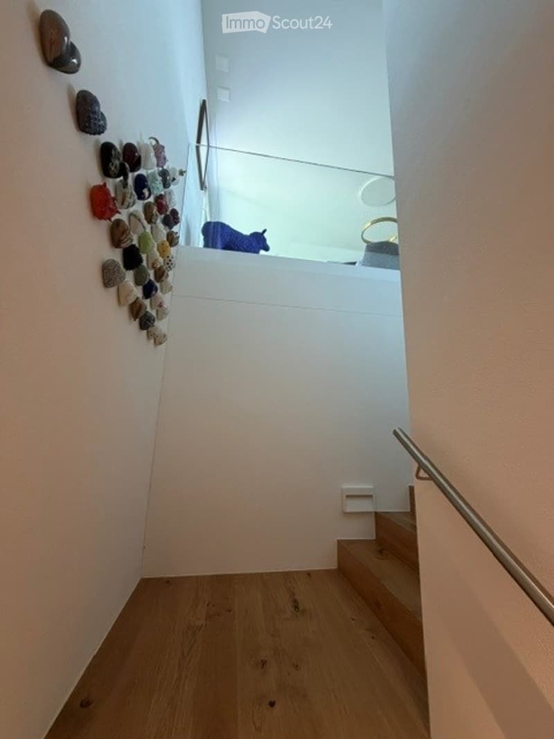 A modern hallway with wooden flooring, white walls, and a staircase leading upwards. A decorative wall features a collection of colorful stones arranged in a pattern. A small blue stuffed animal is visible on the upper landing.