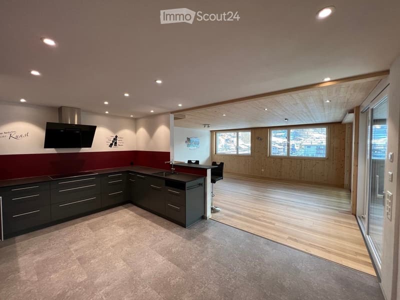 Modern kitchen with dark grey cabinets, a central island, a sink, and a range hood. The space is open to a wooden-floored living area with large windows.