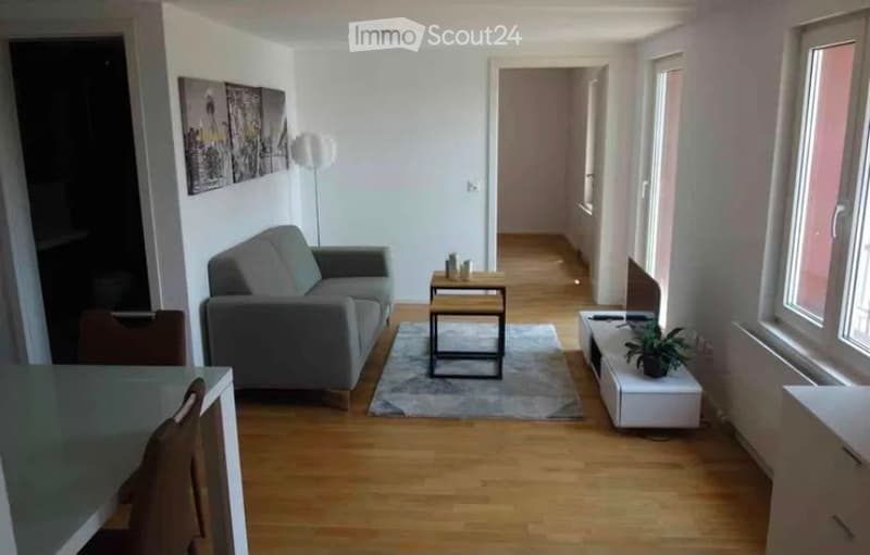 Modern living room with light wooden flooring, grey sofa, coffee table, small side table, and a plant on a white console. The room features large windows and a hallway leading to another area.