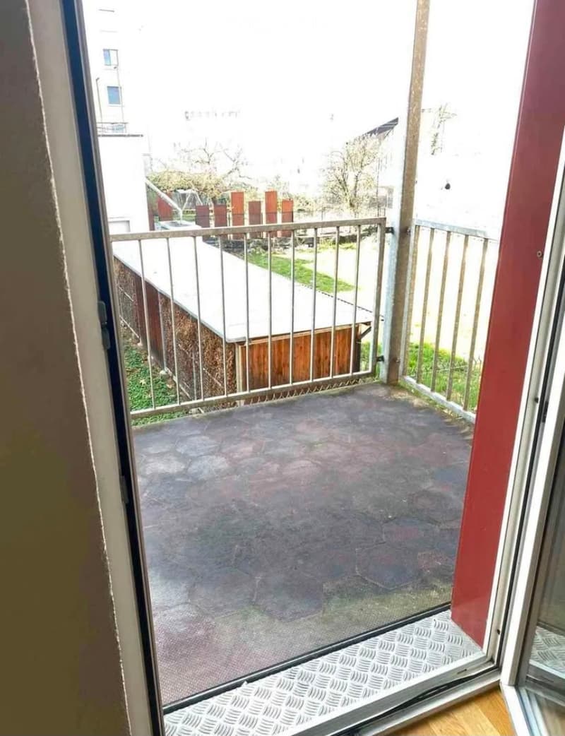 Balcony with metal railing, concrete floor, and view of neighboring buildings and greenery.