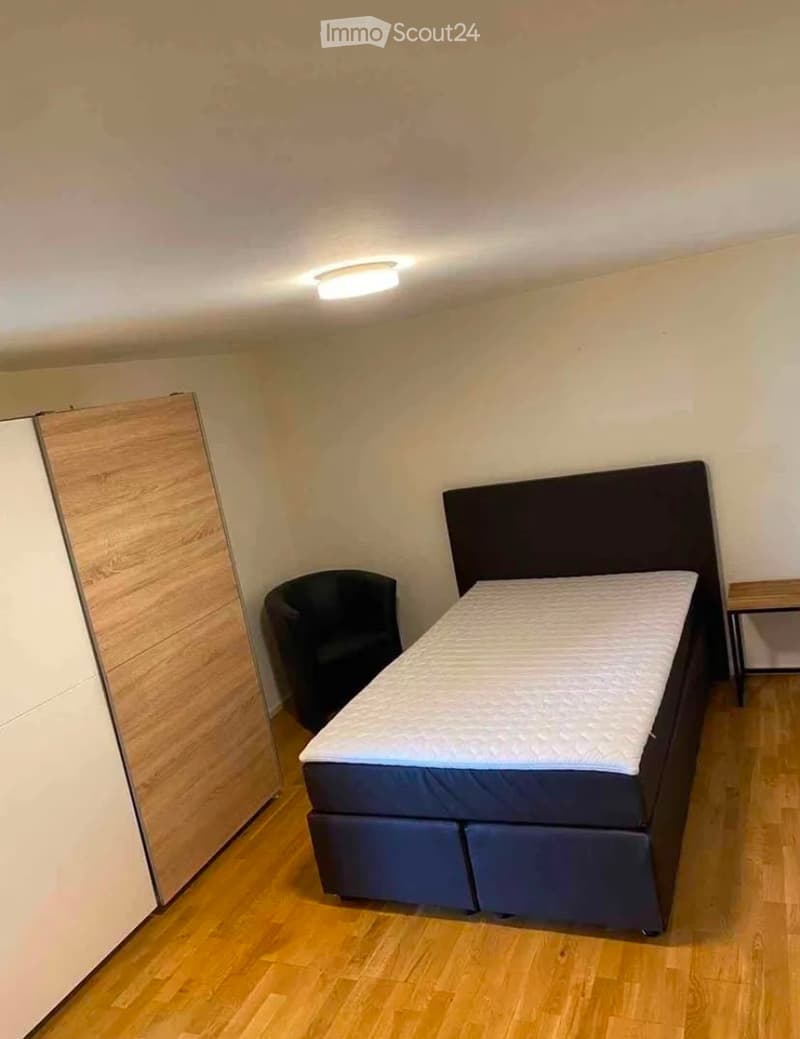 A small, furnished bedroom with a bed, a chair, and a wardrobe. The room has wooden flooring and white walls.