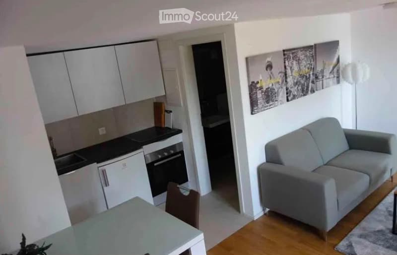 Modern, open-plan living and dining area with a grey sofa, coffee table, and a small dining table with chairs. The kitchen features white cabinets, a stove, and a sink. The walls are decorated with picture frames, and a floor lamp is visible.
