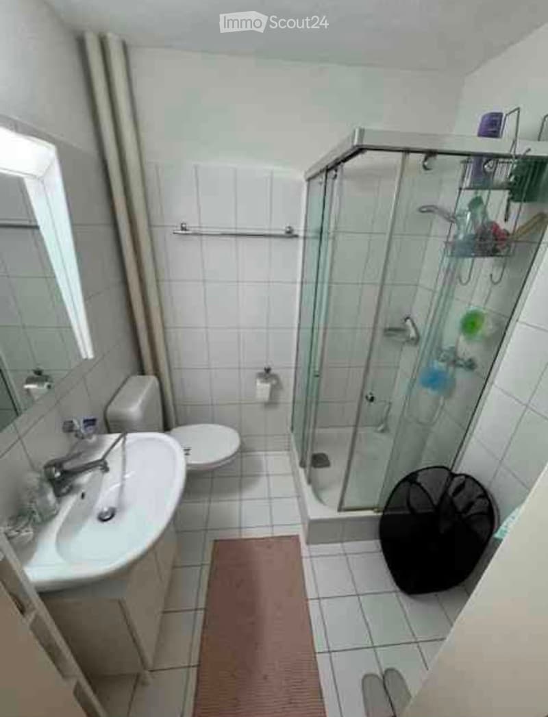 A compact bathroom with white tiled walls and floor, featuring a sink, toilet, and a glass-enclosed shower with a built-in shelf. A small red bath mat is placed in front of the shower.