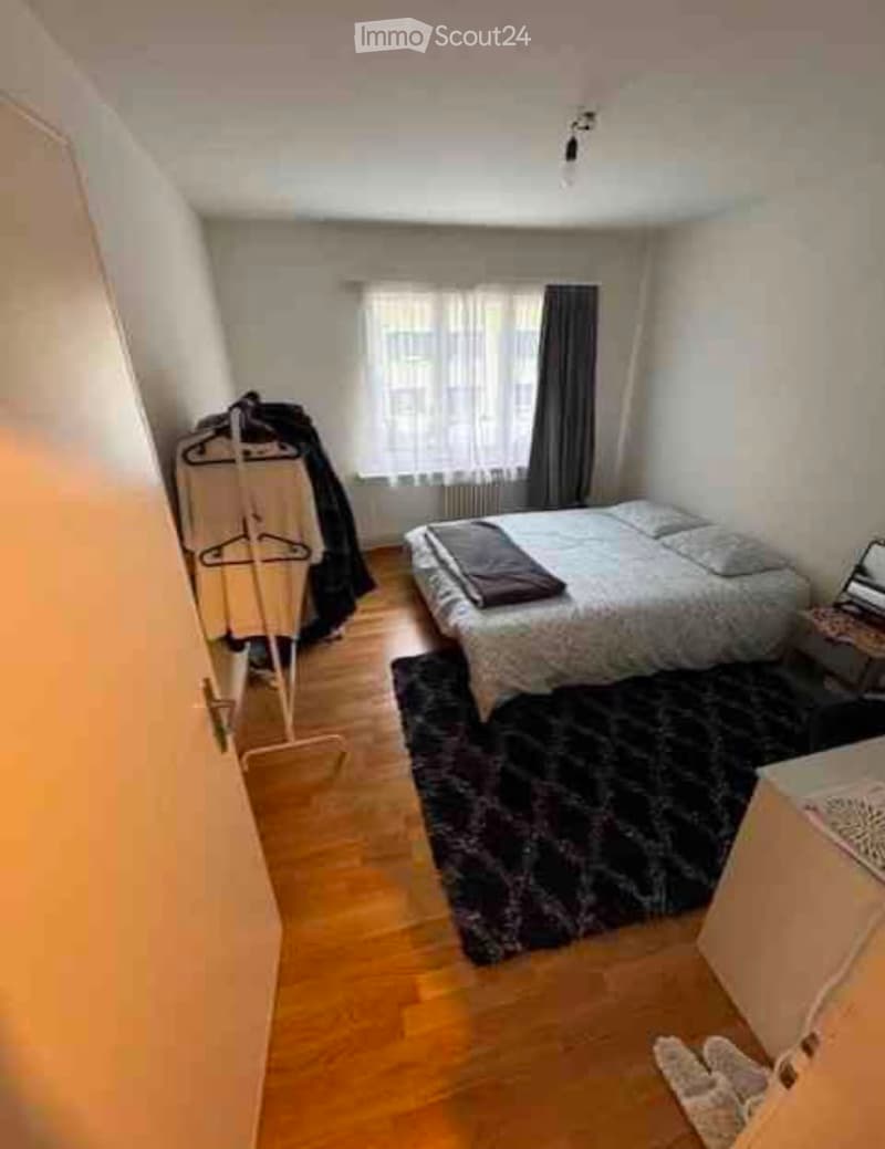 Cozy bedroom with wooden flooring, a double bed with pillows and a blanket, a black rug, a small nightstand, and a clothing rack with hanging clothes.